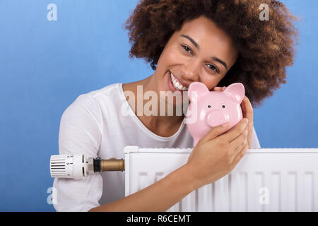 Femme derrière le radiateur de chauffage Holding Piggy Bank Banque D'Images