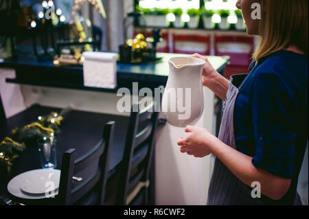 Femme au foyer femme tableau réglage. jeune blonde en Robe et tablier, servi dans une table à manger, plats, effectuent un travail en cuisine. verse le jus d'orange, alcool jaune verseuse tenant dans ses mains. Close-Up Banque D'Images