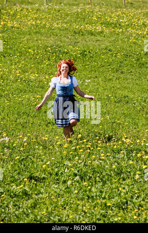 Femme rousse heureux dans le Dirndl, exécutant rire à travers une prairie de fleurs dans les Alpes bavaroises. Banque D'Images