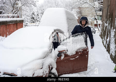 Jeune homme près de voiture couverte de neige. Voitures sous la neige blanche, froide journée d'hiver Banque D'Images