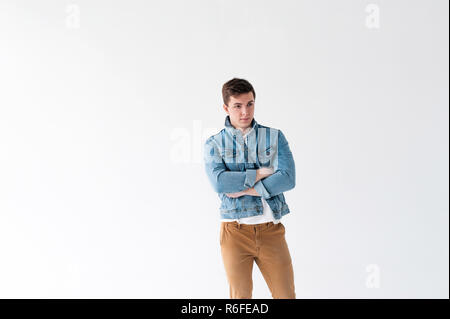 Beau jeune homme de corps du sportif, habillé en veste bleu jeans dans le style de la vieille école et t-shirt blanc avec un pantalon brun, posant sur fond blanc studio, avec les bras croisés Banque D'Images