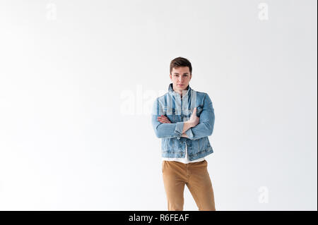 Beau jeune homme de corps du sportif, habillé en veste bleu jeans dans le style de la vieille école et t-shirt blanc avec un pantalon brun, posant sur fond blanc studio, avec les bras croisés Banque D'Images