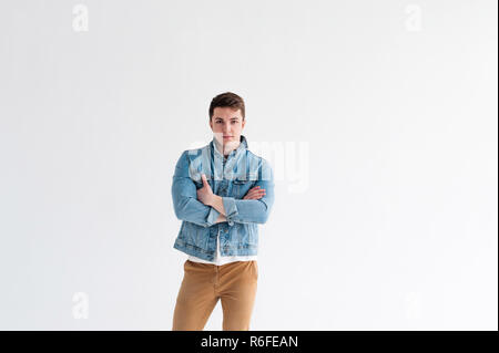 Beau jeune homme de corps du sportif, habillé en veste bleu jeans dans le style de la vieille école et t-shirt blanc avec un pantalon brun, posant sur fond blanc studio, avec les bras croisés Banque D'Images