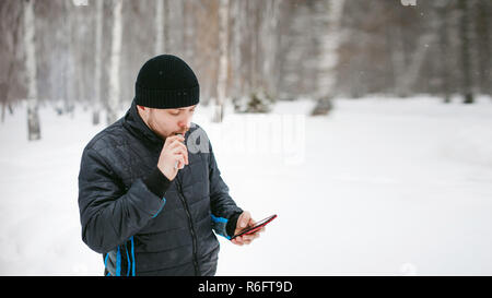 Un jeune homme avec une barbe, fume une cigarette électronique et un plaisir de parler au téléphone par Internet, un effet positif de détente en plein air dans la neige en hiver Banque D'Images