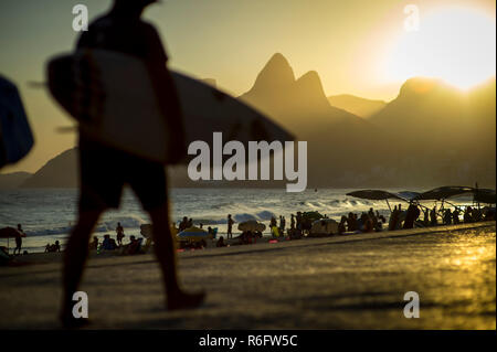 Silhouettes de surfeurs de la marche sur la promenade de la plage d'Ipanema en face d'une vue panoramique sur le soleil derrière deux frères Mountain à Rio Banque D'Images