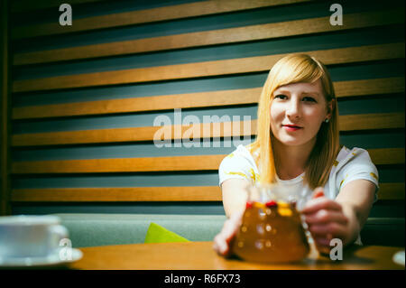 Young blonde woman wearing white T-shirt avec print, girl est titulaire d'une verseuse en verre à la main, assis à une table dans un café, la texture du bois stylisé d'arrière-plan éclairé avec une lumière chaude Banque D'Images