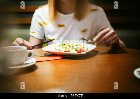 Young blonde woman wearing white T-shirt avec print, fille mange de salade à l'heure du déjeuner, assis à une table dans un café, la texture du bois stylisé d'arrière-plan éclairé avec une lumière chaude Banque D'Images