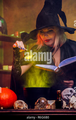 Photo de sorcière blonde en chapeau noir avec le livre de potion de brassage en pot avec vapeur verte Banque D'Images