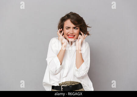 Portrait d'une jeune femme ennuyé habillé en blanc shirt isolés sur fond gris, couvrant les oreilles Banque D'Images