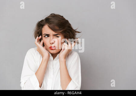 Portrait d'une jeune femme irritée habillé en chemise blanche isolés sur fond gris, couvrant les oreilles Banque D'Images