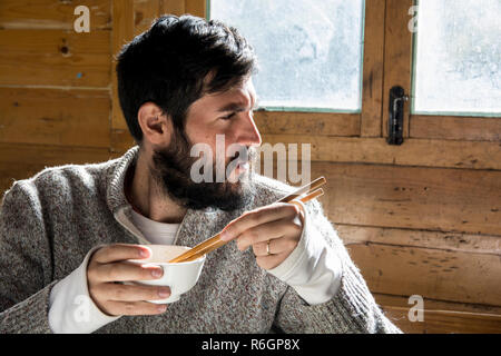 Portrait d'un homme barbu coin et tenait un bol et baguettes dans une cabane en bois, Yading, Chine. Banque D'Images