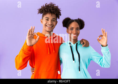 Photo de happy african american couple dans des vêtements colorés montrant ok signer sur fond violet isolés Banque D'Images