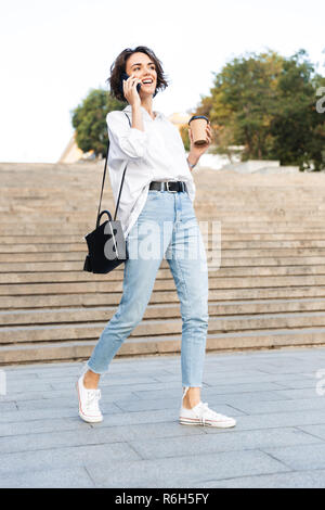 Jolie jeune femme marche dans la rue en bas, talking on mobile phone, boire du café Banque D'Images