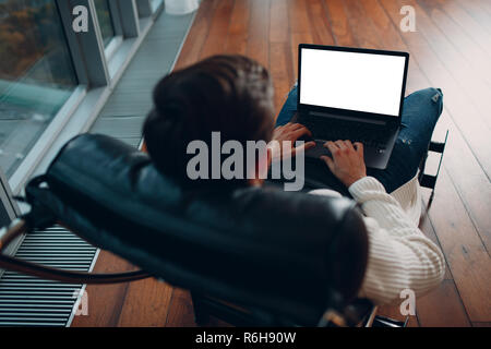 Un jeune homme avec un ordinateur portable avec écran blanc est assis sur une chaise par la fenêtre dans un gratte-ciel. Banque D'Images