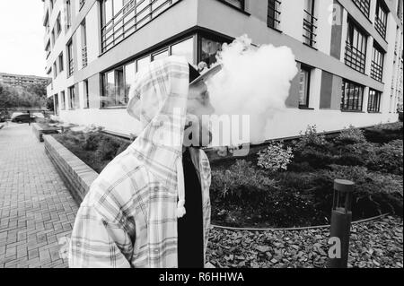 Jeune homme à la barbe en damier lumière veste avec capuche dans un chapeau et des lunettes de marcher librement autour de ville fume un e-cigarette Banque D'Images