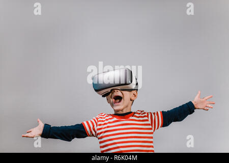Portrait of happy child wearing casque de réalité virtuelle contre un arrière-plan gris. Heureux gamin avec mains propagation s'amusant avec lunettes VR. Banque D'Images