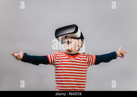 Portrait of happy child wearing casque de réalité virtuelle contre un arrière-plan gris. Heureux gamin avec mains propagation s'amusant avec lunettes VR. Banque D'Images