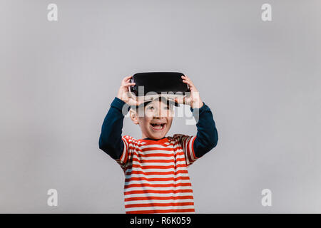 Portrait of cheerful enfant portant casque de réalité virtuelle contre un arrière-plan gris. Smiling happy boy appréciant la réalité virtuelle VR avec lunettes. Banque D'Images