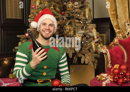 Guy in santa hat sourire avec bouteille de champagne. L'homme en costume elf avec présente des encadrés à arbre de Noël. Donner de cadeau, boxing day. Célébration, party concept. Heureuse nouvelle année, noël, jours fériés. Banque D'Images