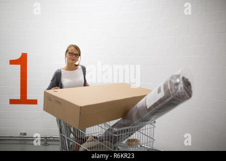 Jolie jeune femme choisir le bon mobilier pour son appartement dans un magasin d'accessoires de maison moderne - avec un chariot, fait du shopping, de quitter le magasin, aller à la maison avec la marchandise achetée(couleur tonique DOF) peu profond de l'image Banque D'Images