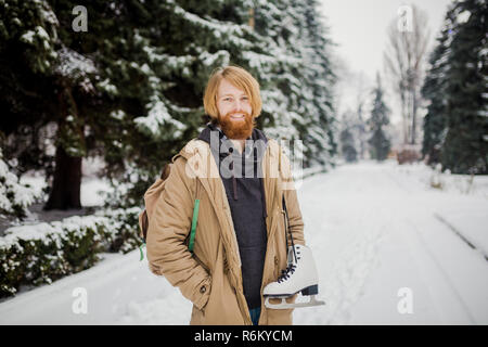 Thème Sports d'hiver. Portrait. Beau jeune homme de race blanche avec de longs cheveux et barbe rouge. En hiver pose modèle snow park contre l'arrière-plan de la forêt sur l'épaule d'une paire de patins en cuir Banque D'Images