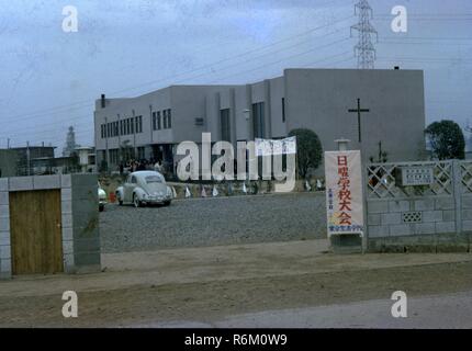 Photo en couleur d'un groupe de Japonais qui se trouvait à l'extérieur, près de l'entrée d'un grand, béton, bâtiment de style Art Déco (peut-être un hôpital ou une église) avec une fenêtre ou bien de la lumière dans la forme d'une croix chrétienne sur une extrémité de l'édifice, un signe écrit en caractères japonais attachés à des perches en bambou s'étendant sur le trottoir une entrée privée, d''une Volkswagen Beetle garée dans un lot, et un sol en bois et béton mur visible dans l'avant-plan, probablement photographié au Japon au cours de la moitié du xxe siècle, 1965. () Banque D'Images