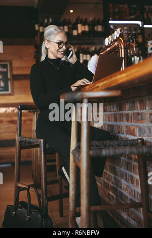 Portrait de femme senior heureuse parlant au téléphone tout en travaillant sur ordinateur portable. femme d'affaires travaillant dans un café. Banque D'Images