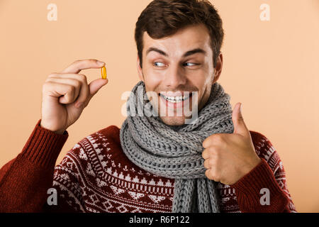 Portrait d'un homme habillé en chandail et isolé sur fond beige écharpe, montrant comprimés capsules, giving Thumbs up Banque D'Images