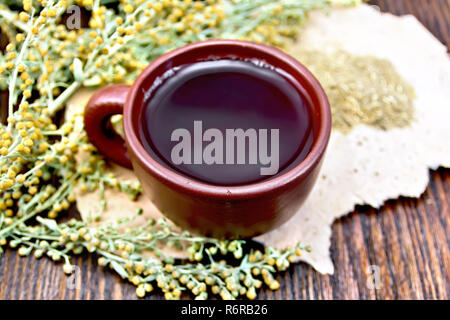 Tisane dans une tasse d'argile brun gris, des brindilles, de l'armoise absinthe séché sur du papier rugueux sur une planche en bois foncé Banque D'Images