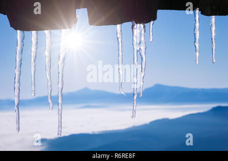 Fond d'hiver avec les glaçons sur le toit d'une maison en bois. Décongeler dans un village de montagne. Beau temps avec ciel bleu Banque D'Images