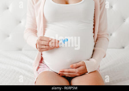 Libre shot of white Caucasian pregnant woman belly avec mignon petit nouveau-né sucette. S'attendant femme femme assise sur lit dans la chambre sucette holding Banque D'Images