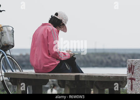 Femelle adulte avec képi siège au banc en bois et lit un livre au parc au bord de mer, à côté de son vélo, Hualien, Taïwan. Tons Vintage Banque D'Images