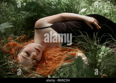 Femme aux cheveux rouges romantique allongé dans l'herbe dans les bois. Une fille dans une robe noire dort et rêve dans une forêt magique Banque D'Images