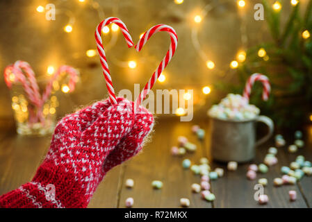 La femme en rouge mitaines tricotées coeur fait hollding à partir de cannes de bonbon. Lumières en effet bokeh, tasse avec de la guimauve sur l'arrière-plan. Concept de carte de Noël, Banque D'Images