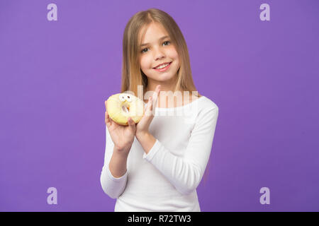 Voulez-vous une morsure ? Happy kid avec de la malbouffe. Sourire de l'enfant avec donut sur fond violet. Petite fille avec bague donut glacé sur fond violet. La nourriture et le dessert. L'enfance et de l'enfants. Banque D'Images