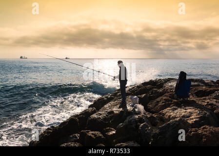 Un pêcheur debout avec la canne à pêche dans la main, il est sur les roches au coucher du soleil, près de lui des seaux et de l'équipement dans des sacs, sur le fond d'un réservoir d'huile Banque D'Images