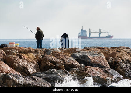 Un pêcheur debout avec la canne à pêche dans la main, il est sur les roches alors qu'il regarde autour de lui, près de lui des seaux et des équipements dans les sacs, sur Banque D'Images