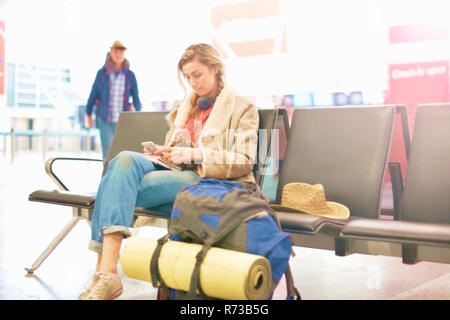 Jeune femme à l'aéroport, assis avec sac à dos à côté d'elle, using smartphone Banque D'Images