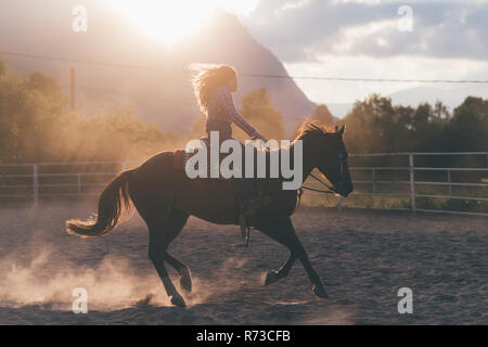 Jeune femme aux cheveux longs au galop sur l'arène équestre en milieu rural, rétroéclairé, Primaluna, Trentino-Alto Adige, Italie Banque D'Images