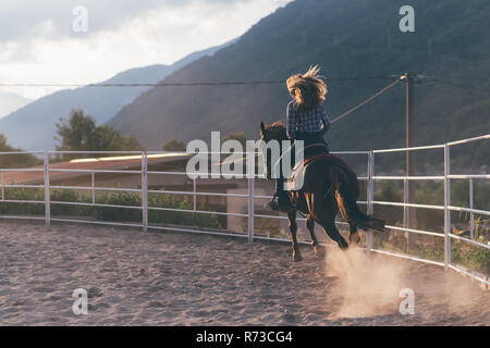 Jeune femme aux cheveux longs au galop sur l'arène équestre en milieu rural, vue arrière, Primaluna, Trentino-Alto Adige, Italie Banque D'Images