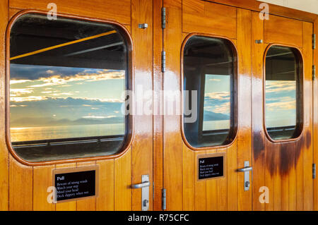 Vernis en bois lourds portes extérieures avec windows et réflexions de mer et soleil du matin dans des panneaux de verre sur un bateau de croisière. L'Alaska, USA. Banque D'Images