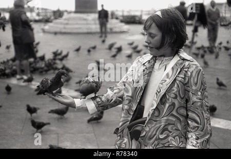 Années 1970, deux pigeons sur le bras d'une jeune fille dans un manteau fleuri à motif debout sur la Piazza San Marco (Sqaure de Saint Marc) Venise, Italie. Les pigeons ont toujours été une attraction touristique populaire à Venise et autrefois rivalisaient avec les chats comme les mascottes traditionnelles, même si non officielles, de la ville de la lagune. Banque D'Images