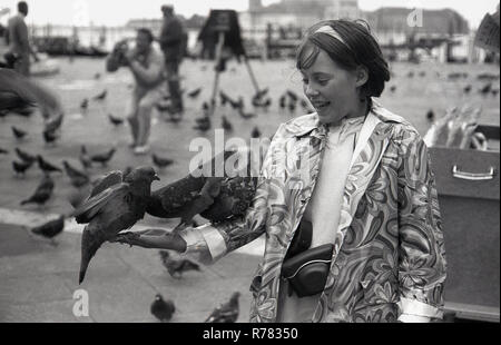 Années 1970, pigeons sur le bras d'une jeune fille en manteau à motifs aflavers debout sur la Piazza San Marco ou la place Saint-Marc, Venise, Italie. Les pigeons ont toujours été une attraction touristique populaire à Venise et autrefois rivalisaient avec les chats comme les mascottes traditionnelles, même si non officielles, de la ville de la lagune. Banque D'Images