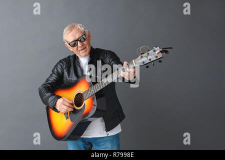 Man en blouson de cuir et lunettes isolés sur gris à la guitare qu'à la joyeuse chantant Banque D'Images