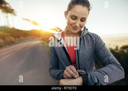 Smiling woman remise en forme contrôle de temps au cours de sa promenade matinale avec le soleil en arrière-plan. Portrait d'une coureuse de régler sa montre tout en jo Banque D'Images