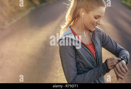 femme de fitness regardant sa montre-bracelet pendant sa course de fitness du matin. Athlète féminine souriante vérifiant le temps debout sur la route tout en écoutant du musi Banque D'Images