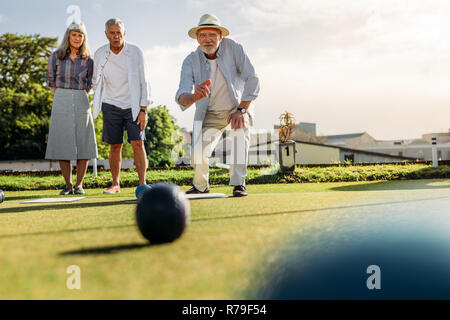 Senior man in hat jeter une tranche alors que ses amis. Deux vieux hommes et une femme jouant un jeu de boules dans une pelouse sur une journée ensoleillée. Banque D'Images