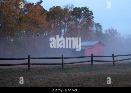 Une vieille grange rouge derrière une clôture en bois sur un matin brumeux juste avant le lever du soleil. Lake Benson Park à Garner la Caroline du Nord. Banque D'Images