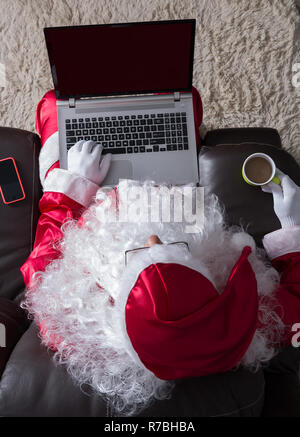 Passage du Père Noël (ou Kris Kringle) se détendre dans un canapé à la maison. En utilisant un ordinateur portable pour la communication et les loisirs et de boire du café avec du lait. Banque D'Images