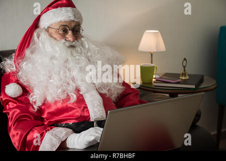 Sur le côté du Père Noël (ou Kris Kringle) se détendre dans un canapé à la maison à l'aide d'un ordinateur portable pour la communication et les loisirs ou les achats en ligne. Banque D'Images